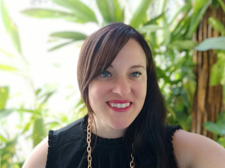 Photo of Kim McDaniels smiling, wearing a black sleeveless top with a gold necklace. There are tropical plants in the background.